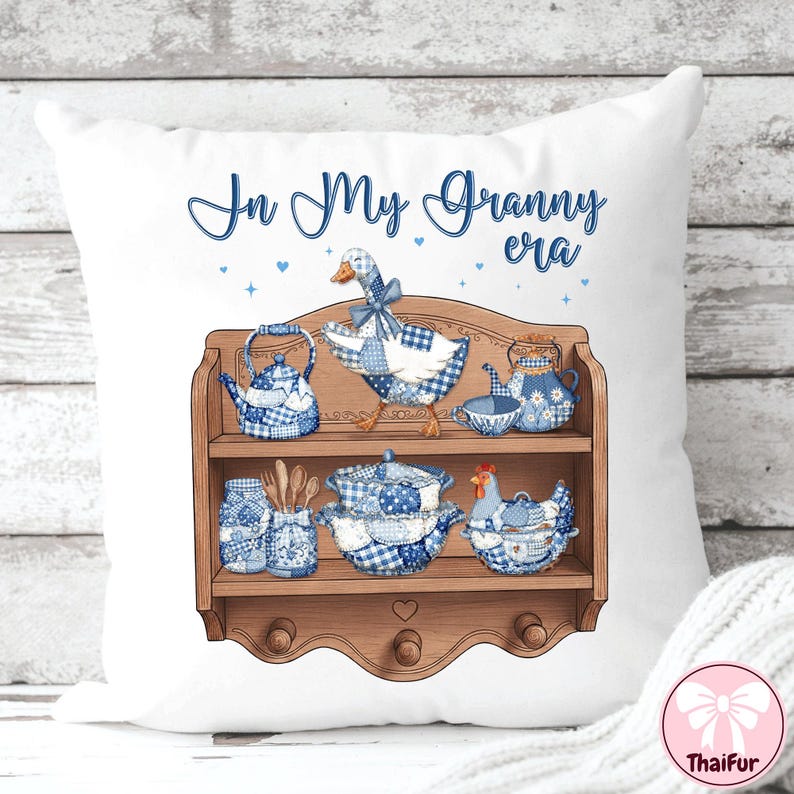 K&ouml;nnte beinhalten: Wei&szlig;es Zierkissen mit einem blau-wei&szlig; karierten G&auml;nse- und K&uuml;chenutensilien-Design. Der Text "In My Granny era" ist in blauer Schrift. Das Design umfasst ein Holzregal mit Teekannen, einem Huhn und anderen K&uuml;chenartikeln.