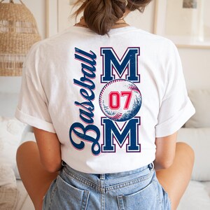 May include: White t-shirt with a baseball-themed design. The design features the word "MOM" with a baseball graphic and the number "07" in the center. The word "Baseball" is written vertically on the left side of the design.