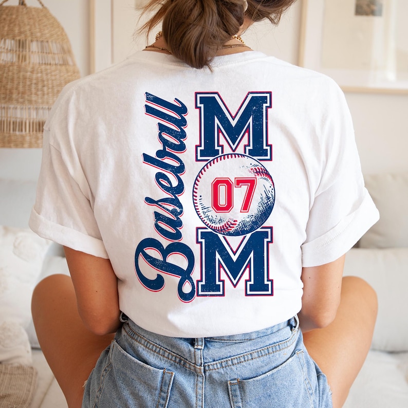 May include: White t-shirt with a baseball-themed design. The design features the word "MOM" with a baseball graphic and the number "07" in the center. The word "Baseball" is written vertically on the left side of the design.