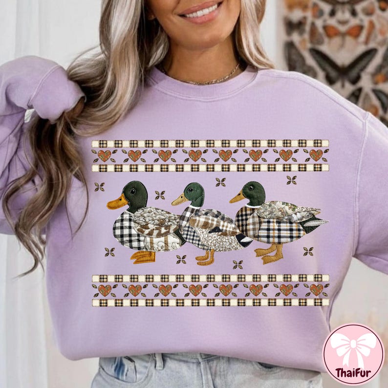 May include: A light purple sweatshirt featuring three ducks with plaid patterns and green heads. The ducks are adorned with brown and white plaid patterns. The design includes decorative borders with hearts and squares.