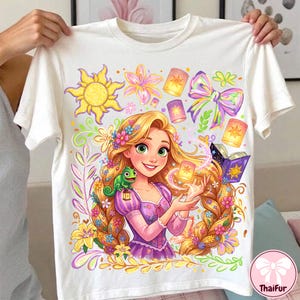 Puede incluir: Una camiseta blanca de manga corta con un estampado gráfico colorido de un personaje de pelo largo con un camaleón verde, un emblema de sol, linternas flotantes, un libro morado y detalles florales, con un gran lazo iridiscente en la parte superior.