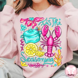 May include: Pink sweatshirt with a colorful graphic design. The design includes a lobster, lemon slices, a cooking pot with the word "Summer", and the phrase "'Tis The Seasoning". The design is in shades of pink, blue, yellow, and green.