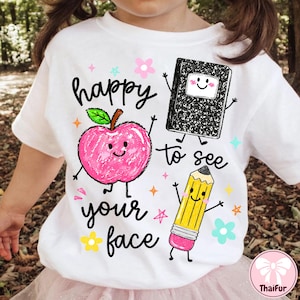 May include: A white t-shirt featuring a colorful graphic print of a pink apple, a black composition notebook, and a yellow pencil, all with smiling faces. The text "happy to see your face" is printed in black script alongside small stars and flowers.