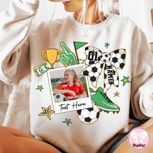 May include: White sweatshirt with a green, yellow, and black soccer-themed design. The design includes a soccer ball, a trophy, a hand pointing, a soccer cleat, a bow, and a photo frame with a girl holding a soccer ball. The text "Text Here" is printed on the photo frame. The bow has the text "NAME" and "01" printed on it.