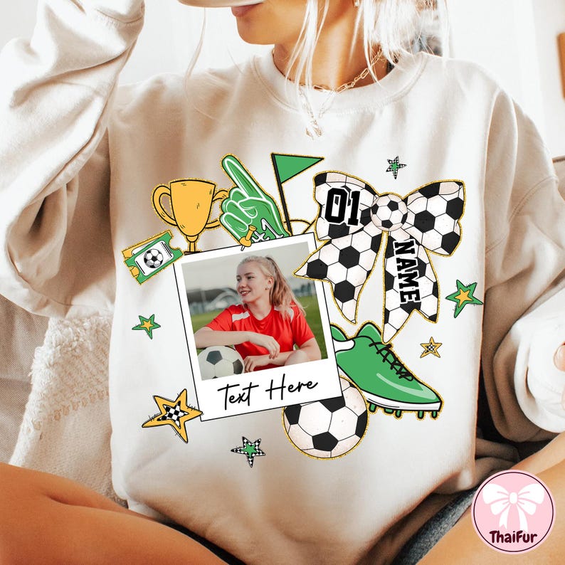 May include: White sweatshirt with a green, yellow, and black soccer-themed design. The design includes a soccer ball, a trophy, a hand pointing, a soccer cleat, a bow, and a photo frame with a girl holding a soccer ball. The text "Text Here" is printed on the photo frame. The bow has the text "NAME" and "01" printed on it.