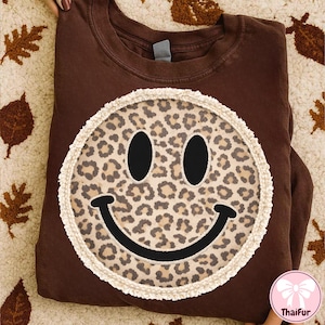May include: A brown sweatshirt featuring a large smiley face with a leopard print design. The smiley face has black eyes and a black smile, surrounded by a cream-colored trim. The sweatshirt is folded and displayed on a patterned surface.
