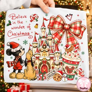 Puede incluir: Un cuadrado de tela blanca con una colorida ilustración navideña que muestra un castillo, Mickey Mouse, Pluto, un gran lazo a cuadros y una taza. El texto dice Believe in the wonder of Christmas y merry CHRISTMAS con detalles festivos.