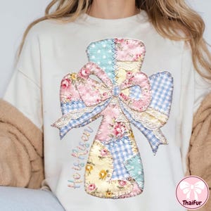 Op de afbeelding: Crèmekleurige sweatshirt met een patchwork kruisontwerp en een grote strik. Het kruis is gemaakt van bloemen-, stippen- en ruitjesstoffen, met de woorden "He is Risen" aan de zijkant geborduurd. Het ontwerp bevat kanten afwerking.