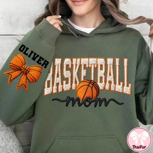 Puede incluir: Una sudadera con capucha verde oliva con las palabras "BASKETBALL mom" en bordado blanco y negro, y un gr&aacute;fico de baloncesto. La manga izquierda presenta el nombre "OLIVER" y un lazo naranja.