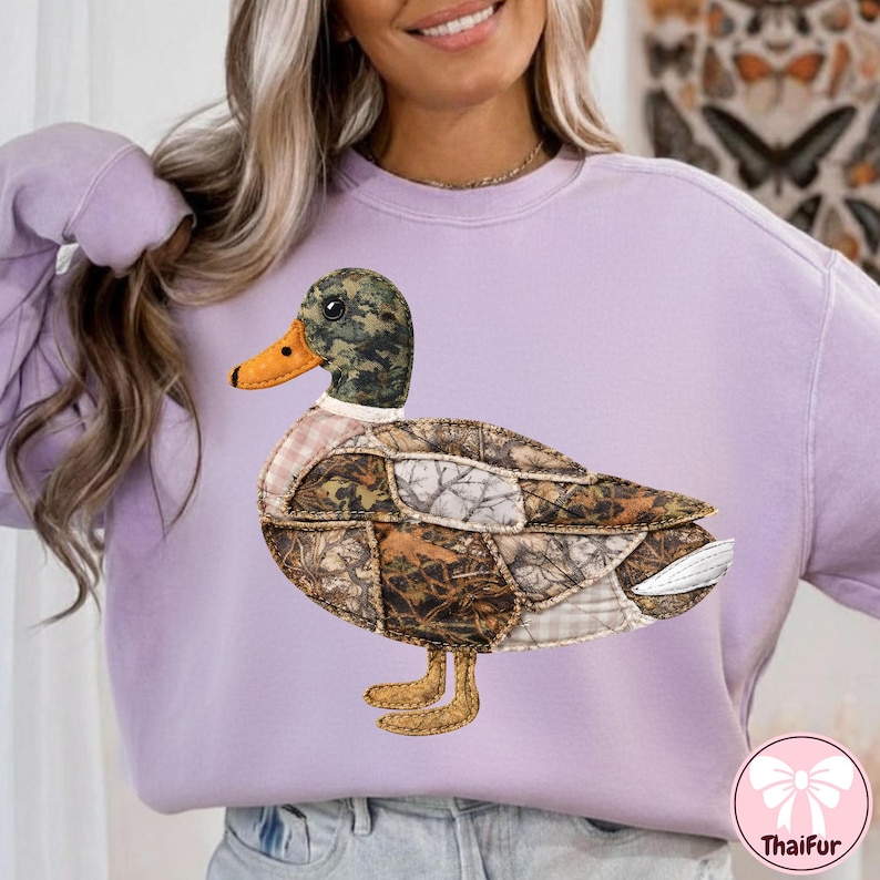May include: A light purple sweatshirt featuring a detailed appliqu&eacute; of a duck. The duck has a camouflage head, a patchwork body with various earth-tone fabrics, and an orange beak. The duck's feet are yellow.