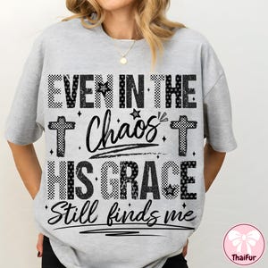 May include: A light gray t-shirt with the text "EVEN IN THE Chaos HIS GRACE Still finds me" in a mix of fonts and patterns, including checkered and script styles. The design includes cross symbols and stars.
