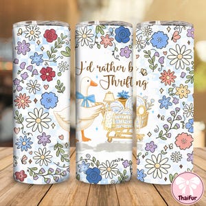 May include: A white tumbler with a floral design and the text "I'd rather be Thrifting." The tumbler features a cartoon goose with a blue bow, a shopping cart filled with dishes, and a light blue checkered background.
