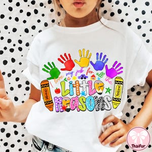 May include: White t-shirt featuring a colorful design with handprints and the text "I love my job for all the Little Reasons." The design includes orange and yellow crayons, a car, a house, and a star.