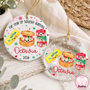 Puede incluir: Dos adornos redondos, uno de cerámica blanca y otro de acrílico transparente, cada uno con un cordón dorado. Ambos muestran el texto THE YEAR OF SQUISHY HUNTING, Octavia y 2026, junto a ilustraciones de mantequilla, un dumpling y tres squishies.