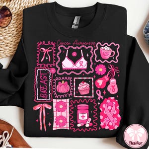 May include: Black sweatshirt with pink graphic squares featuring a bra, ribbon, flowers, and the text "Cancer Awareness" and "BREAST". The design includes a pink ribbon, a cupcake, and a pink pumpkin.