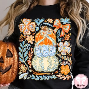 May include: Black sweatshirt featuring a stacked pumpkin design with floral and bow accents. The pumpkins are orange, blue, and cream-colored, with a bow on top. Surrounding the pumpkins are orange and blue flowers and leaves.