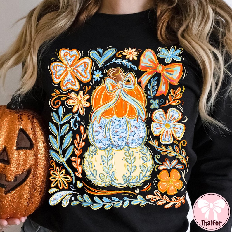 May include: Black sweatshirt featuring a stacked pumpkin design with floral and bow accents. The pumpkins are orange, blue, and cream-colored, with a bow on top. Surrounding the pumpkins are orange and blue flowers and leaves.