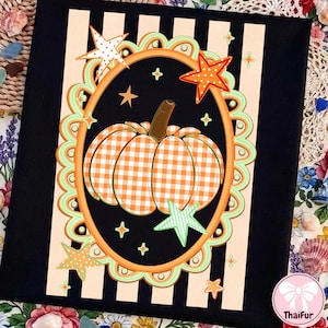 May include: A black fabric panel featuring a central oval frame with a scalloped light green border. Inside the oval is an orange and white gingham pumpkin with a brown stem, surrounded by orange and green stars on a black background with stripes.