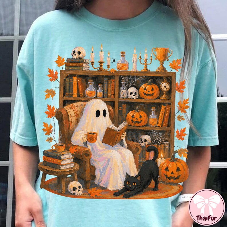 May include: A light blue t-shirt featuring a graphic of a white ghost sitting in an armchair reading a book. The scene includes a bookshelf with pumpkins, skulls, candles, and a black cat. Orange autumn leaves surround the central illustration.