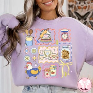 May include: A lilac sweatshirt with a charming, illustrated design of kitchen and baking items. The design includes a chicken, jam jars, a teapot, cookies, and a scale. The overall aesthetic is whimsical and charming, ideal for those who enjoy baking.