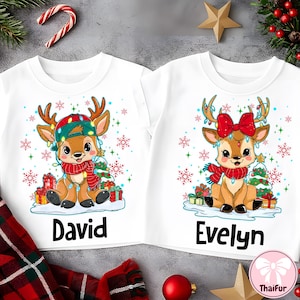 May include: Two white Christmas-themed t-shirts. One features a cartoon reindeer wearing a hat and scarf, with the name "David" below. The other shirt shows a reindeer with a bow and scarf, with the name "Evelyn". Both shirts have snowflake and gift box graphics.