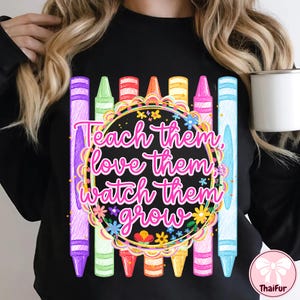 May include: A black long-sleeve sweatshirt featuring a central graphic of colorful crayons arranged vertically behind a circular frame. The text "Teach them, love them, watch them grow" is written in pink script inside the floral-decorated circle.