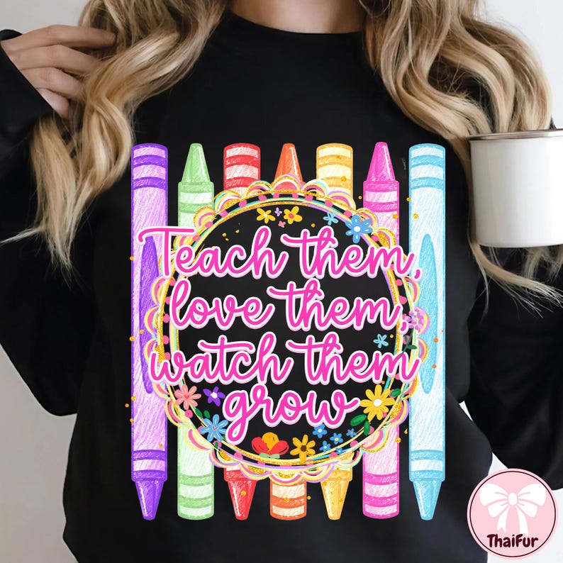 May include: A black long-sleeve sweatshirt featuring a central graphic of colorful crayons arranged vertically behind a circular frame. The text "Teach them, love them, watch them grow" is written in pink script inside the floral-decorated circle.