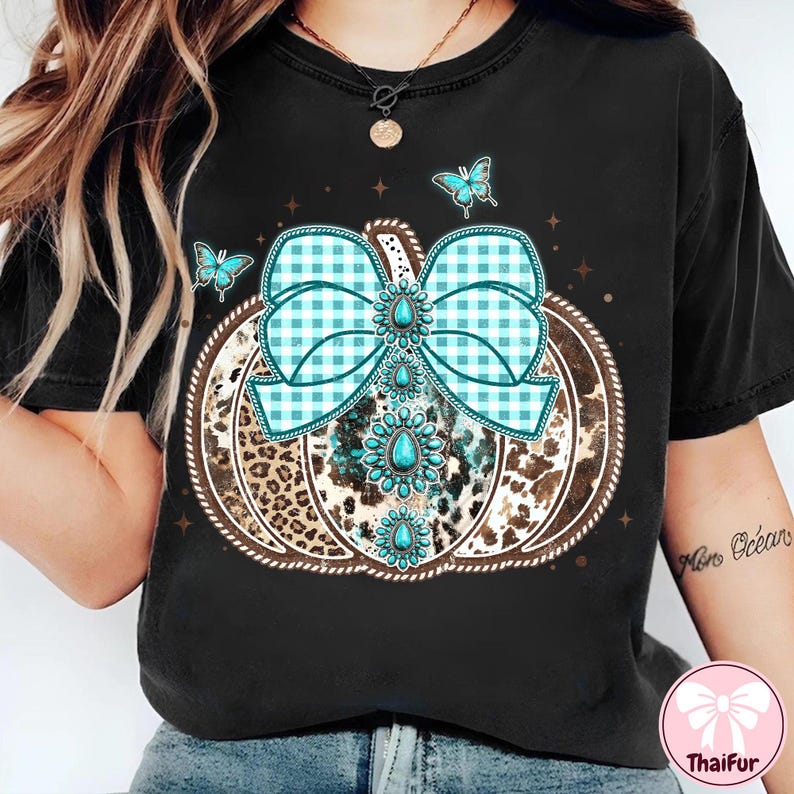 May include: Black t-shirt featuring a pumpkin design with a turquoise and white gingham bow, turquoise gemstones, and a leopard print pattern. The pumpkin is outlined with a brown rope-like border, and there are three turquoise butterflies.