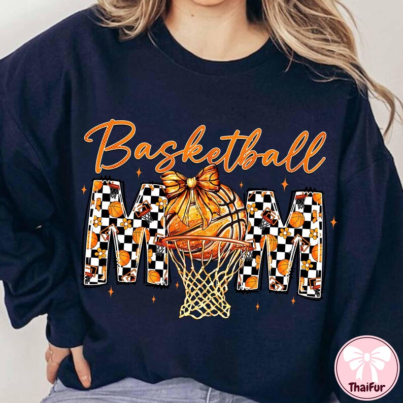 May include: Navy blue sweatshirt with the words "Basketball Mom" in orange script. The design features a basketball with a bow, a net, and the letters "MOM" in a checkered pattern with basketballs and flowers.