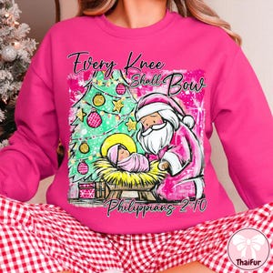 May include: A pink sweatshirt with a Christmas-themed graphic. The design features a Christmas tree, Santa Claus, and a baby in a manger, with the text "Every Knee Shall Bow" and "Philippians 2:10".