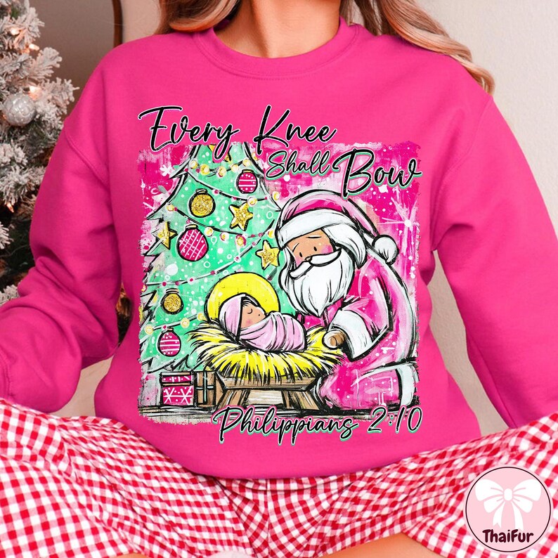 May include: A pink sweatshirt with a Christmas-themed graphic. The design features a Christmas tree, Santa Claus, and a baby in a manger, with the text "Every Knee Shall Bow" and "Philippians 2:10".