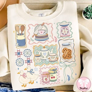 Peut inclure: Sweat-shirt cr&egrave;me avec un motif sur le th&egrave;me de la p&acirc;tisserie. Le graphisme comprend des illustrations d'ustensiles de cuisine, d'une th&eacute;i&egrave;re, de biscuits, de canards et de pots de confiture, le tout encadr&eacute; de bordures d&eacute;coratives. Le design est en couleurs pastel douces.