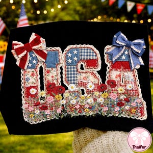 May include: Black fabric with the word "USA" appliqued in red, white, and blue patchwork fabric. The letters are decorated with bows and a floral border with daisies and poppies. The design has a patriotic theme.