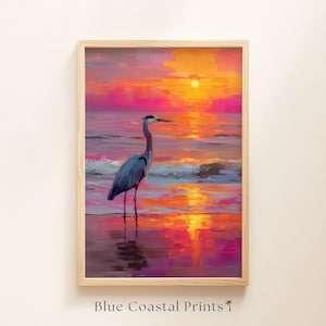 May include: Framed art print of a blue heron standing in shallow water at sunset. The sky is a vibrant mix of pinks, oranges, and yellows, reflecting on the water's surface. The text "Blue Coastal Prints" is at the bottom.
