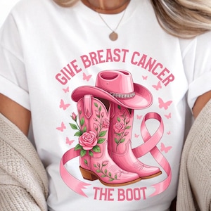 Op de afbeelding: Wit T-shirt met een roze grafisch ontwerp. Het ontwerp toont een roze cowboyhoed en -laarzen, een roze lint en vlinders. De tekst luidt "GIVE BREAST CANCER THE BOOT".