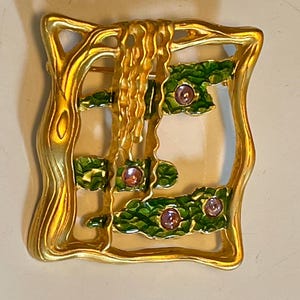 May include: A gold-toned brooch with a green enamel design and pink rhinestones. The brooch features a stylised tree with a flowing design.