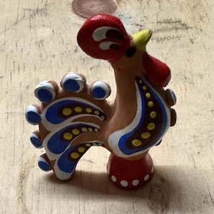 Pode incluir: Uma figura de galo de cerâmica pintada à mão com uma crista vermelha, um bico amarelo, penas de cauda azuis e brancas e um corpo castanho com bolinhas brancas e amarelas.