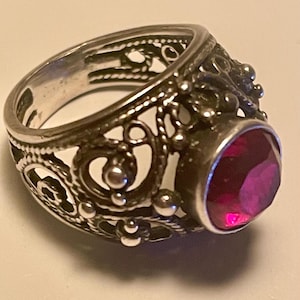 May include: Silver ring with an elaborate filigree design and a large, faceted, oval-shaped, deep red gemstone. The ring has intricate openwork patterns and small, raised details around the band and the gem setting.