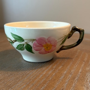 May include: Cream-coloured teacup with a pink floral design and green leaves. The handle is dark green. The teacup is made of ceramic and is likely vintage.