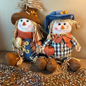 May include: Two plush scarecrow dolls with fabric faces, straw hair, and felt hats. One wears a brown hat and blue overalls, holding corn. The other wears a blue hat and a black-and-white checkered shirt, holding a small orange pumpkin.