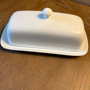 May include: A white rectangular butter dish with a lid and a small, round knob. The dish sits on a matching rectangular base. The butter dish is made of ceramic or porcelain and is designed for storing butter.