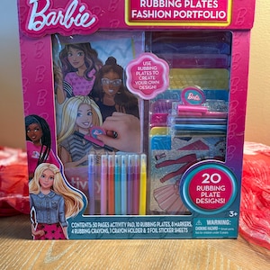 May include: A pink Barbie Rubbing Plates Fashion Portfolio box set. The set includes 50 activity pages, 10 rubbing plates, 8 markers, 4 crayons, a crayon holder, and 2 foil sticker sheets. The box features images of Barbie and the text "Rubbing Plates Fashion Portfolio".
