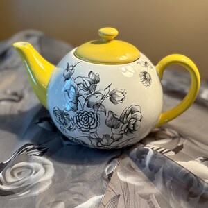 May include: A white ceramic teapot with a yellow lid, handle, and spout. The teapot is decorated with black floral illustrations. The teapot is sitting on a gray floral patterned surface.