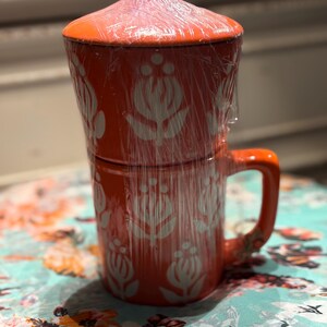 May include: An orange ceramic mug with a matching lid, wrapped in clear plastic. The mug features a white floral pattern. The handle is curved. The mug is sitting on a floral patterned surface.