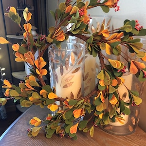 May include: A decorative wreath with green and orange leaves and small red berries. The wreath is placed in front of two glass candle holders with lit candles inside. The candle holders have a leaf pattern.
