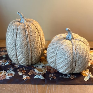 May include: Two decorative pumpkins crafted from tan herringbone fabric, each featuring a prominent vertical jute twine accent and a silver-toned stem. The pumpkins are positioned side-by-side, showcasing a textured weave pattern.