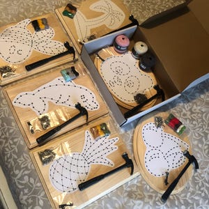 May include: A set of six string art kits featuring various designs, including a pineapple, a dolphin, a palm tree, a heart, and two other designs. Each kit includes a wooden board, nails, yarn, and a hammer.
