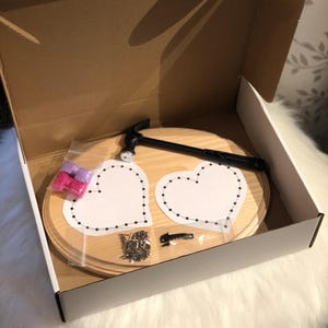 May include: A craft kit in an open cardboard box. The kit includes a wooden base, two heart-shaped templates, a hammer, nails, and pink and purple thread. The wooden base is light brown, and the templates are white.