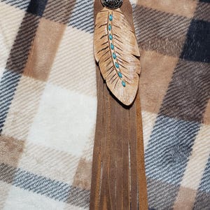 May include: A brown leather keychain with a feather design. The feather is adorned with turquoise-colored accents and a silver-toned metal button. The keychain has a fringe of leather strips and a silver clasp.