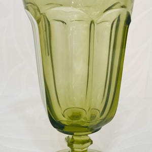 May include: A vintage-style, olive green glass goblet. The goblet features a flared rim, a faceted stem, and a round base. Vertical lines run down the sides of the glass, adding to its classic design. The glass is transparent, allowing light to pass through.